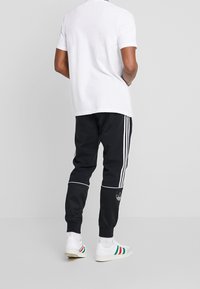 Man som bär vita sneakers med röda och gröna ränder, svarta träningsbyxor med vita ränder, och en vit kortärmad skjorta, står vänd bort.