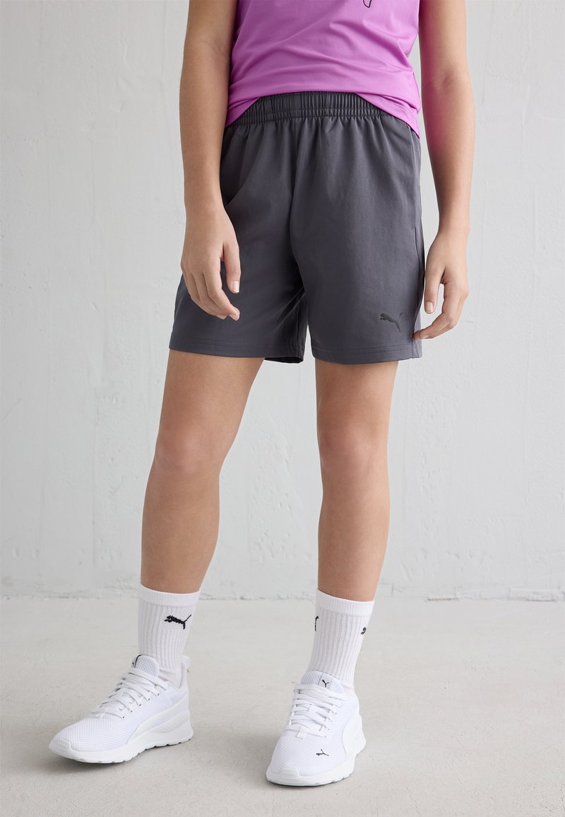 Isik, kes kannab tumehalli Puma lühikesi pükse, valgeid Puma sokke ja valgeid sportjalatseid, seisab ühtlast valget seina ja põranda taustal.