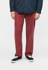 Άτομο που φοράει κόκκινο παντελόνι κορντόρι, μαύρα και λευκά αθλητικά παπούτσια και σκούρο μπλε πουκάμισο με κουμπιά, με τα χέρια χαλαρά στο πλάι.