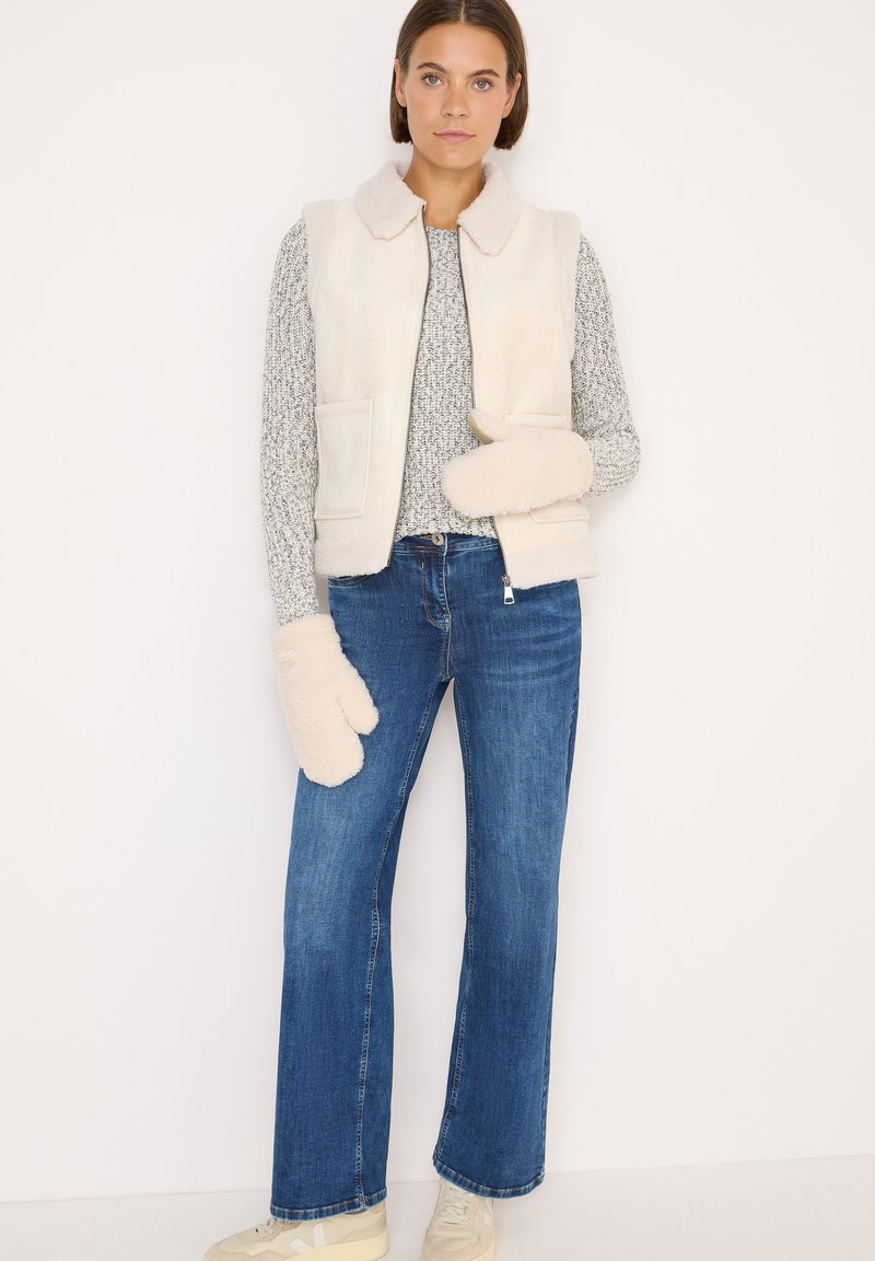 Hørfrakk i off-white med krave, matchende en grå mønstret sweater, blå vidde jeans og cremefarvede vanter.