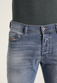 Modré džínové kalhoty mají klasický design s pěti kapsami, vybledlé nohavice, jemné naznačené šisování a zapínání na kovový knoflík a zip.