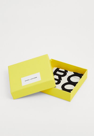 Dzeltena taisnstūra kastīte ar baltu etiķeti, kurā rakstīts "MARC JACOBS." Iekšā ir rakstains audums ar drosmīgiem melniem burtiem uz balta fona.