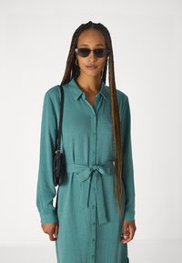 Abito lungo color teal con bottoni e cintura annodata, realizzato in un tessuto texture. Borsa a spalla nera e occhiali da sole scuri completano l'outfit.