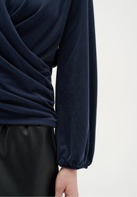 Femme portant un haut bleu marine à manches longues avec un détail de couture froncée et une jupe en cuir noir, sur un fond clair uni.