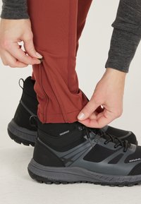 Vattentäta svarta vandringsskor i kombination med rödbruna byxor med dragkedjor vid anklarna, mot en neutral bakgrund.