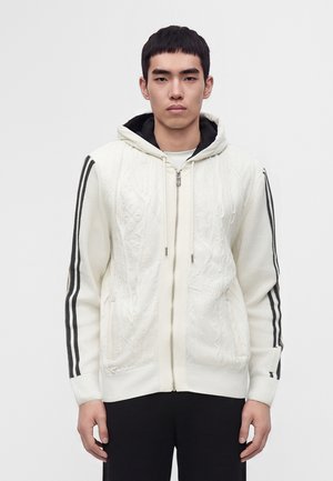 Young man wearing a white zip-up knitted hoodie with black stripes on sleeves and black pants, standing against a plain light background.