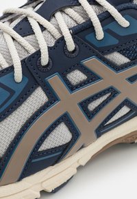 Running shoe with a grey mesh upper, navy blue synthetic overlays, beige accents, and white laces. Textured sole for traction.