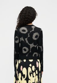 Μαύρη πλεκτή ζακέτα με ανθισμένο μοτίβο, ριμπ υφή και στρογγυλή λαιμόκοψη, συνδυασμένη με κίτρινη πλισέ φούστα με αφηρημένα σχέδια.