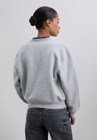 Sudadera gris con cuello redondo y hombros caídos. Tejido suave, puños y bajo acanalados, con un cuello interior oscuro a contraste.