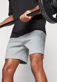 Persona che indossa pantaloncini grigi e maglietta nera solleva un bilanciere con dischi di peso su uno sfondo semplice.