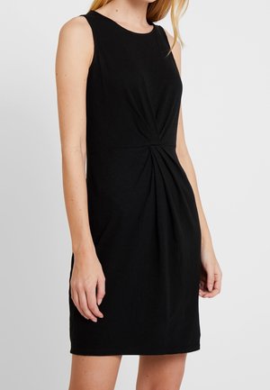 Ärmelloses schwarzes Kleid mit gerafftem Stoffdetail an der Taille, getragen von einer stehenden Person mit heller Haut und langen blonden Haaren.