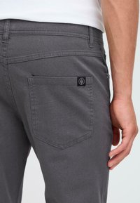 Grijze spijkerbroek met een achterzak met een contrasterende zwarte logo patch. De stof heeft een zachte textuur en traditionele stikseldetails.