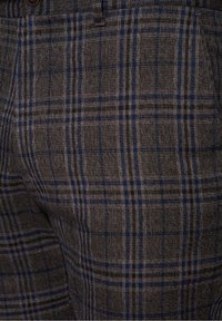 Zbliżenie na materiał w brązowo-niebieską kratę na dopasowanych spodniach z widoczną kieszenią i guzikiem w pasie.