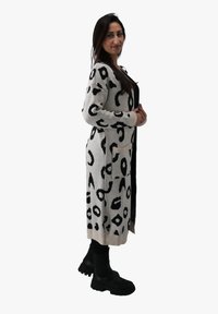 Lungo cardigan color crema con stampa leopardata nera, dotato di tasche laterali, polsini a coste e una vestibilità rilassata. Indossato sopra pantaloni neri e stivali.