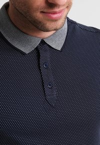 T-shirt polo marine avec un motif de pois blancs, dotée d'un col texturé contrastant, d'une patte de boutonnage à trois boutons et de manches courtes.