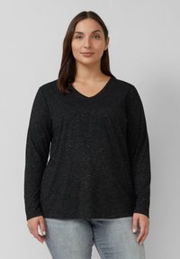 Frau mit braunen Haaren, die ein schwarzes langärmliges V-Ausschnitt-Shirt und hellblaue Jeans trägt, steht vor einem schlichten grauen Hintergrund.