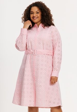 Lächelnde Frau mit lockigem Haar, die ein pinkfarbenes, mit Gürtel versehenes Kleid mit Lochstickerei und Blumendekor sowie langen Ärmeln trägt, vor einem einfarbigen Hintergrund.