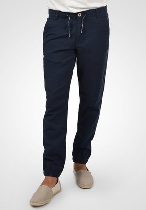 Marineblaue Jogginghosen aus Baumwolle mit einem elastischen Bund, Kordelzug und Bündchen an den Knöcheln. Es werden hellbeige Slipper getragen.