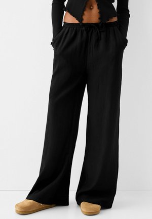 Persoon die losse zwarte broek draagt met handen in de zakken, een cropped zwart topje en beige instapschoenen, staand tegen een witte achtergrond.