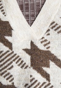 Gros plan sur un pull tricoté à col en V, affichant des motifs géométriques et étoilés marron et crème sur un tissu texturé.