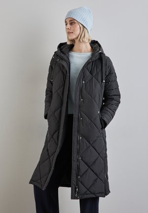 Gequilted zwarte lange jas met een kap, voorzien van drukknoopsluitingen en zijzakken. Vergezeld van een lichtblauwe geribbelde gebreide beanie.