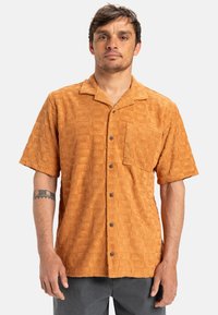 Short-sleeve, terry cloth shirt in burnt orange with a textured pattern of squares and sunburst designs; single chest pocket and black buttons.