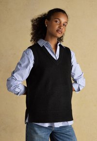 Frau mit lockigem Haar, die einen schwarzen ärmellosen Pullover über einem blau-weiß gestreiften Hemd und blauen Jeans trägt, steht mit den Händen hinter dem Rücken.