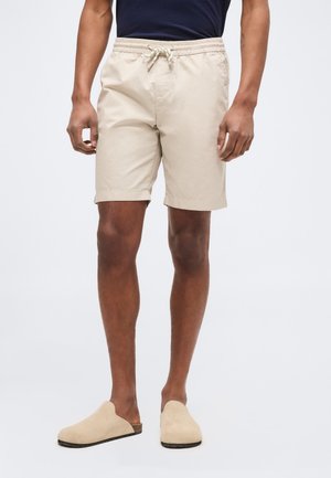 Mann trägt beige Shorts mit Kordelzug, navyblaues Hemd und beige Slipper, steht vor einem schlichten hellen Hintergrund.