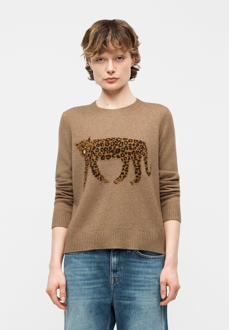 Jeune personne aux cheveux courts et ondulés portant un pull marron avec un motif léopard brun foncé et un jean bleu, debout devant un fond uni.