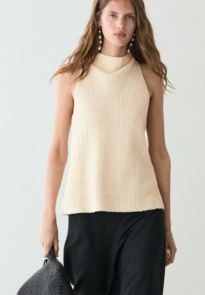 Donna con capelli lunghi indossa un top beige senza maniche in maglia e pantaloni neri, tenendo una borsa nera intrecciata su uno sfondo chiaro e semplice.