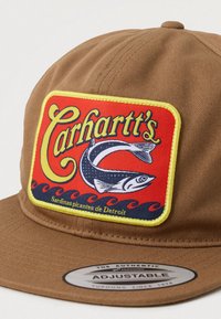 Cască ajustabilă maro cu o pată roșie rectangulară, pe care se află textul galben "Carhartt's", o ilustrație cu pește și textul "Sardinas picantes de Detroit."