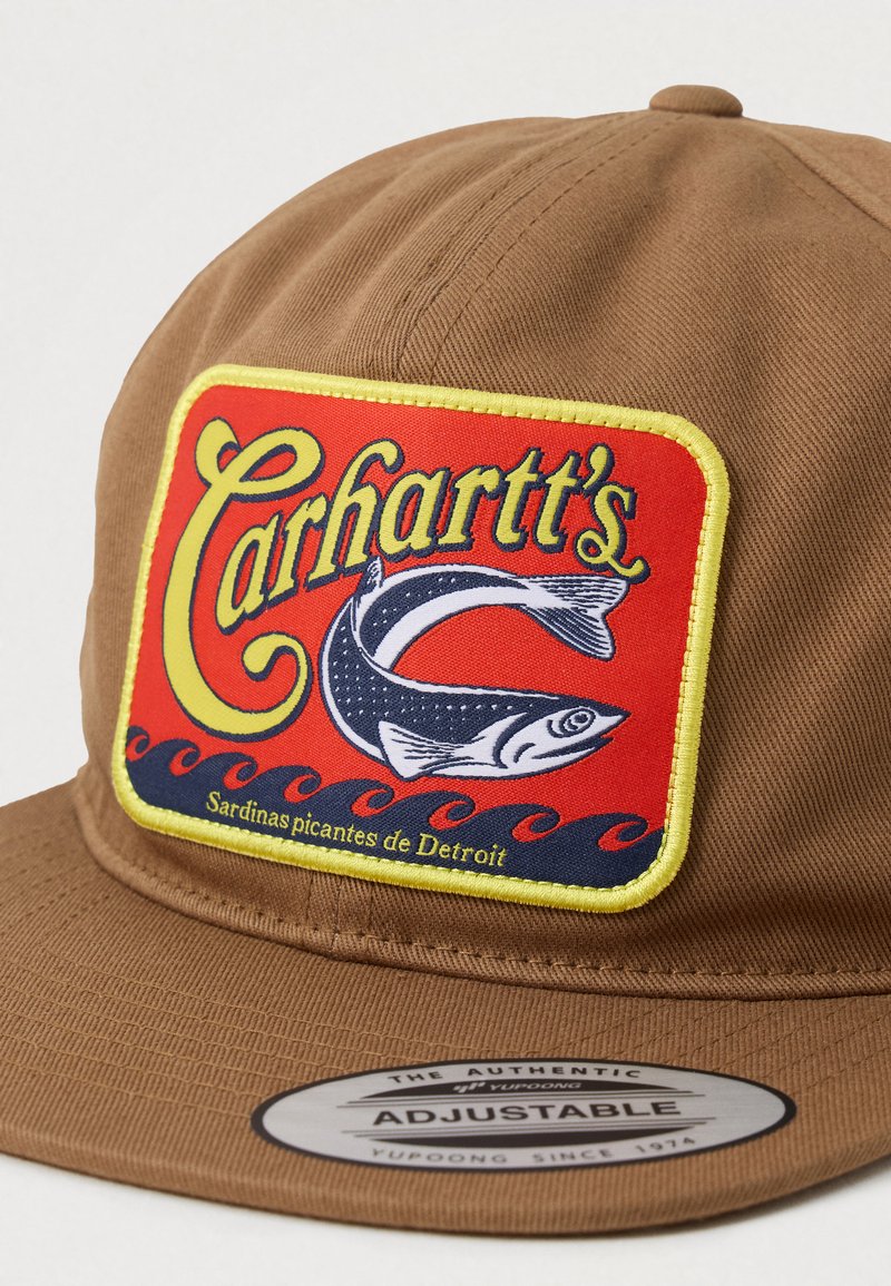 Braune verstellbare Kappe mit einem rechteckigen roten Patch, der gelbe "Carhartt's"-Schriftzüge, eine Fischillustration und "Sardinas picantes de Detroit" zeigt.