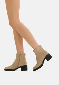 Stivali ankle in suede beige con tacco in gomma nera, zip laterale e un accento dorato del logo sul retro. Texture liscia e design semplice.