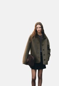 Cappotto in lana oversize verde oliva con colletto alto, chiusura con bottoni e bottoni neri; abbinato a una gonna nera e una borsa marrone.