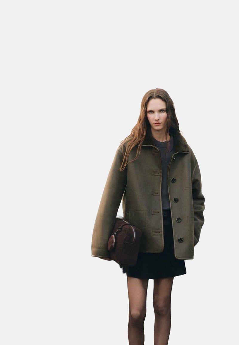 Cappotto in lana oversize verde oliva con colletto alto, chiusura con bottoni e bottoni neri; abbinato a una gonna nera e una borsa marrone.