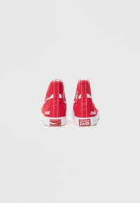 Raudonos aukštos viršutinės dalies sportbačiai iš drobės su baltomis gumos padais. Ypatybės: Coca-Cola ir Converse logotipai, kontrastingos siūlės ir užtrauktukas.