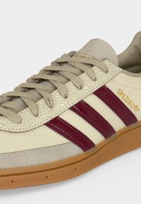Μπεζ αθλητικό παπούτσι Adidas Spezial με μπορντό ρίγες, ταμπ κορδόνια, σουέτ μύτη και σόλα από καουτσούκ, που δείχνει υφή σκισμένο δέρμα.