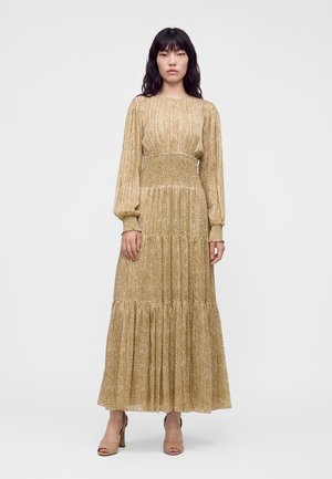 Kvinde med langt sort hår iført en lang, gulvmønstret kjole med lange ærmer og beige åbne sko, stående mod en ensfarvet hvid baggrund.