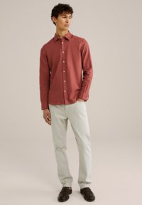 Cămașă roșie cu mâneci lungi și nasturi, confecționată dintr-un material ușor, asociată cu pantaloni de culori deschise și pantofi loafers negri, stând în fața unui fundal neutru.
