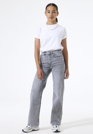 T-shirt blanc à manches courtes associé à un jean en denim gris taille haute. Le mannequin porte des baskets blanches et un collier simple en perles.