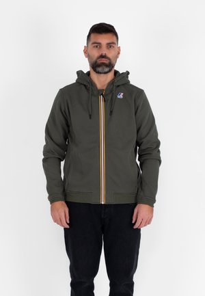 Felpa con cappuccio verde con cerniera e striscia gialla e blu che corre lungo il centro, con cappuccio e logo sul petto. Indossata con jeans scuri.