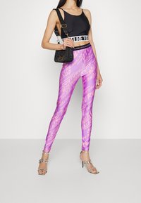 Svart kort tanktopp med ett vitt band och lila leggings med ett glansigt textmönster, kombinerat med strappy silverklackar och en svart väska.