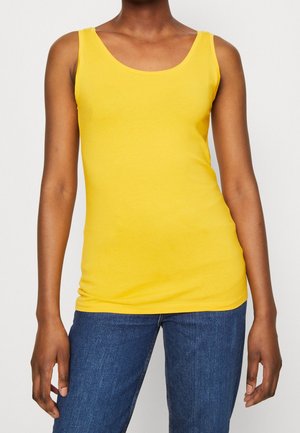 Person, die ein leuchtend gelbes ärmelloses Oberteil und blaue Jeans trägt, steht mit entspannten Armen seitlich an einem einfarbigen Hintergrund.