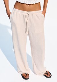 Pantalons à large jambe en ribbed beige clair, avec une taille élastique et un cordon de serrage. Comprend des poches latérales et tombe lâchement sur des tongs noires.