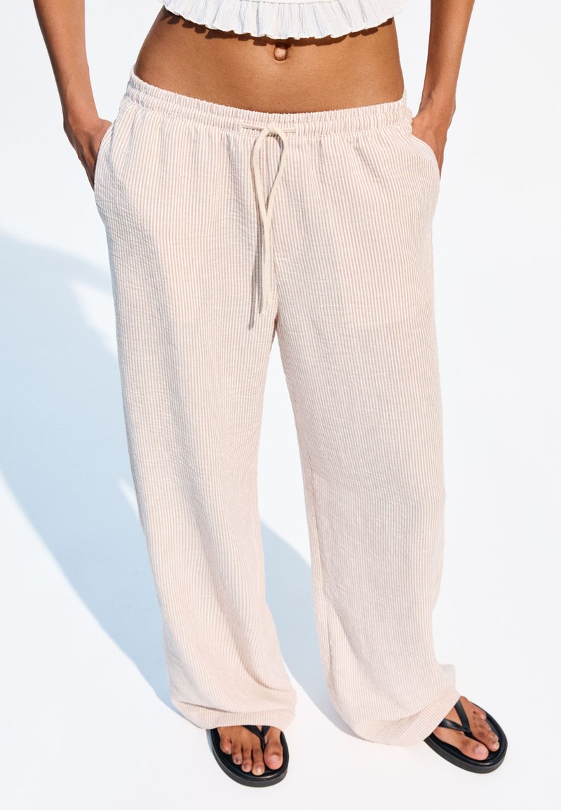 Pantalons à large jambe en ribbed beige clair, avec une taille élastique et un cordon de serrage. Comprend des poches latérales et tombe lâchement sur des tongs noires.
