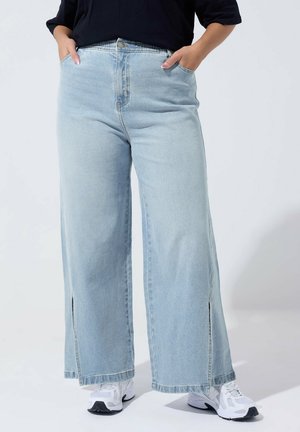 Person iført lyseblå vidde jeans med sidelommer og hvide sneakers, hænderne afslappet i lommerne, foran ensfarvet baggrund.