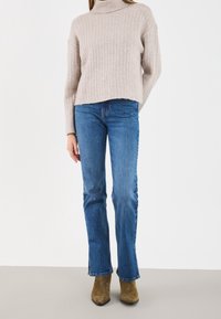 Pull à col roulé beige côtelé, coupe courte ; jean bleu évasé ; bottines en daim marron ; textures lisses ; coutures minimales ; lignes épurées.