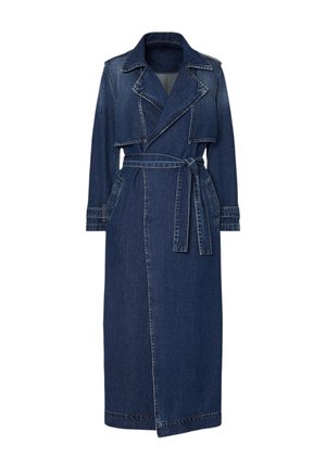 Donna con lungo trench coat in denim blu scuro, cintura legata sul retro, mani nelle tasche e grandi orecchini a cerchio, vista di lato.