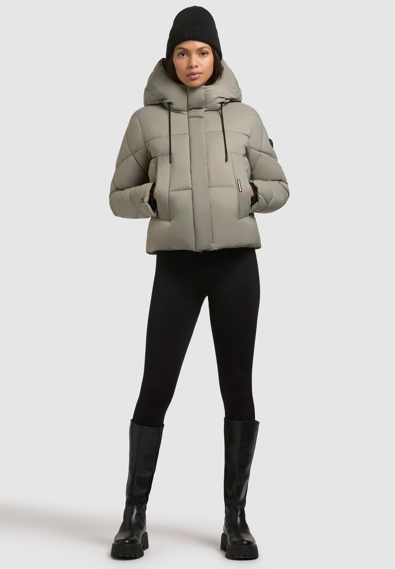 Kvinde iført en lys grå dunjakke med hætte, sorte leggings, sort hue og sorte knæhøje støvler, stående på en grå baggrund.