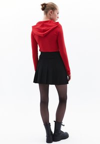 Červený žebrovaný crop top s dlouhým rukávem a kapucí, spárovaný s černou plisovanou mini sukni, černými punčochami a masivními černými šněrovacími botami.